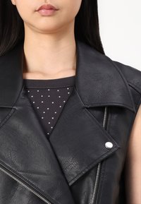 Černá vesta z umělé kůže se širokým klopami a stříbrnými kovovými doplňky. Pod ní tmavý vršek zdobený malými bílými puntíky.