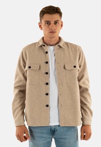 Jonge man in een beige knoopjack met zwarte knopen over een wit T-shirt en blauwe spijkerbroek, staande tegen een effen achtergrond.
