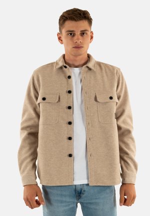Jonge man in een beige knoopjack met zwarte knopen over een wit T-shirt en blauwe spijkerbroek, staande tegen een effen achtergrond.