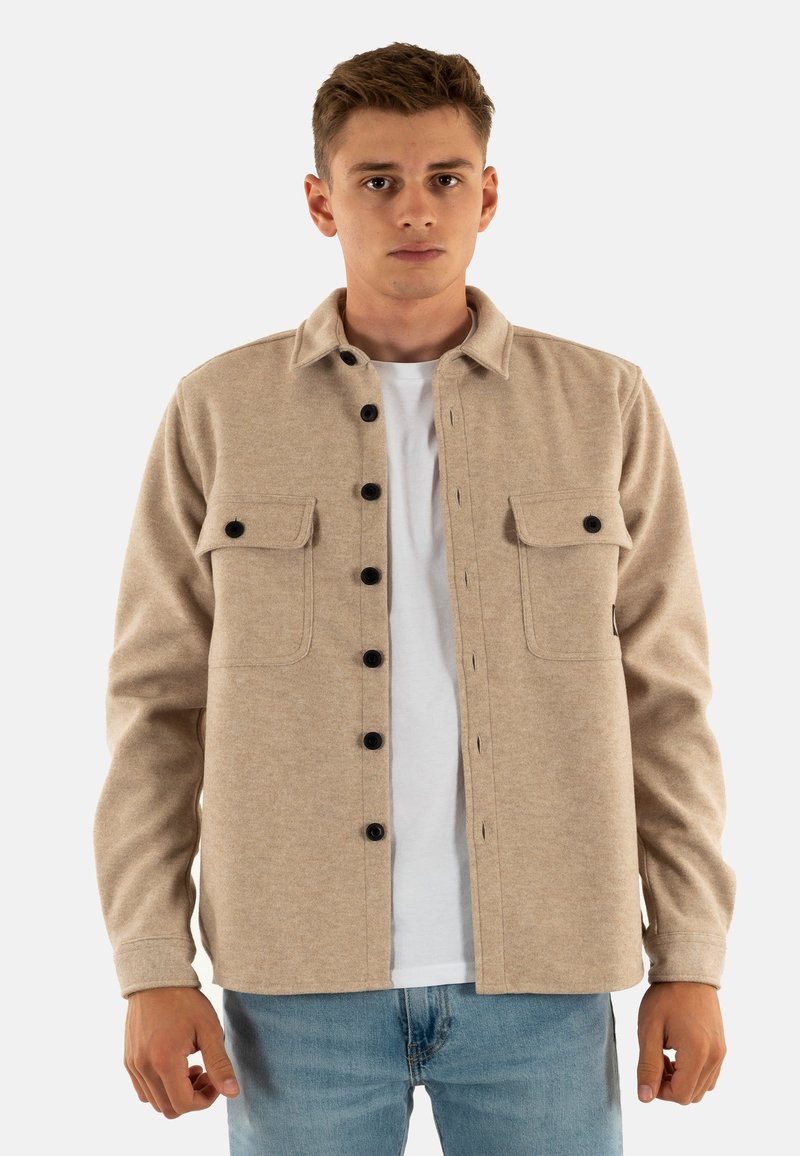 Jonge man in een beige knoopjack met zwarte knopen over een wit T-shirt en blauwe spijkerbroek, staande tegen een effen achtergrond.