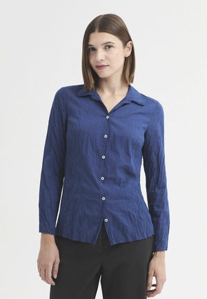 Femme aux cheveux bruns lisses porte une chemise bleue boutonnée à manches longues et un pantalon noir, se tenant devant un fond clair uni.