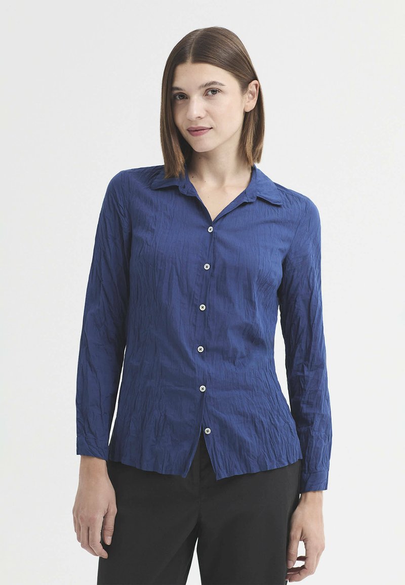 Femme aux cheveux bruns lisses porte une chemise bleue boutonnée à manches longues et un pantalon noir, se tenant devant un fond clair uni.