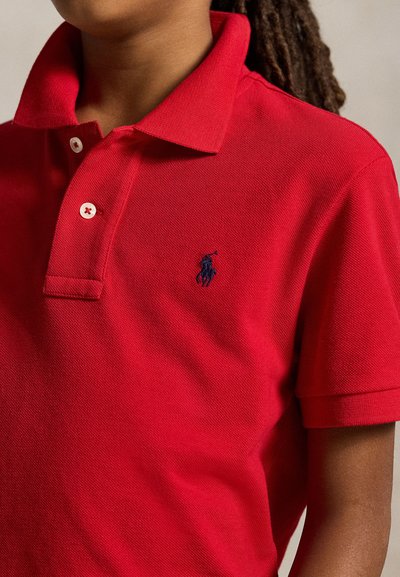 Kind draagt een rood polo met korte mouwen, witte knopen en een klein donker geborduurd paardenlogo op de borst.