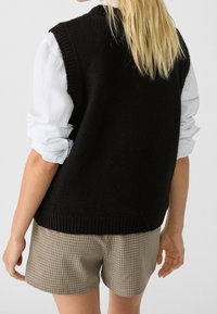 Gilet en laine noir avec un col et un ourlet côtelés, porté par-dessus une chemise bleu clair. En dessous, un short marron à motifs avec un design à carreaux.
