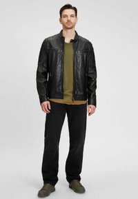 Zwarte leren jas met een ritssluiting, twee borstzakken en gemêleerde schouderaccenten, gedragen over een olijfgroen shirt en zwarte broek.