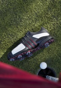Black and white golf shoe with red accents on green grass near a golf hole with a white ball about to drop in.