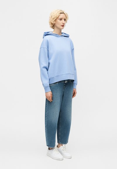 Νεαρός με κοντά κατσαρά ξανθά μαλλιά που φοράει ανοιχτό μπλε φούτερ με κουκούλα, άνετο μπλε τζιν και λευκά αθλητικά, στέκεται πλαγιαστά.