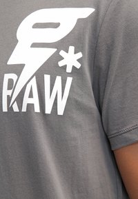Šedé bavlněné tričko s výrazným bílým grafickým designem obsahujícím text "RAW" a stylizované logo. Krátké rukávy a hladká textura.