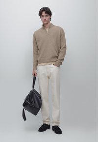 Beige strikket pullover med halvlukket krave, kombineret med cremefarvede bukser og sorte sneakers; holder en sort lædertaske. Minimalistisk design.