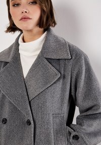 Donna che indossa un cappotto di lana grigio doppiopetto sopra un maglione a collo alto bianco testurizzato, con capelli corti castani e trucco neutro.