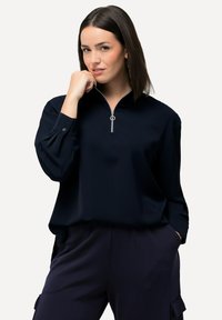 Marineblaue Zip-Up-Top mit langen Ärmeln, aus glattem Material gefertigt. Hat einen hohen Kragen und ein silbernes Reißverschlussdetail am Hals.