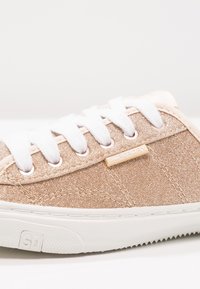 Zapatilla brillante en rosa dorado con cordones blancos, parte superior texturizada, suela de goma y un pequeño parche de logo beige en el lado.