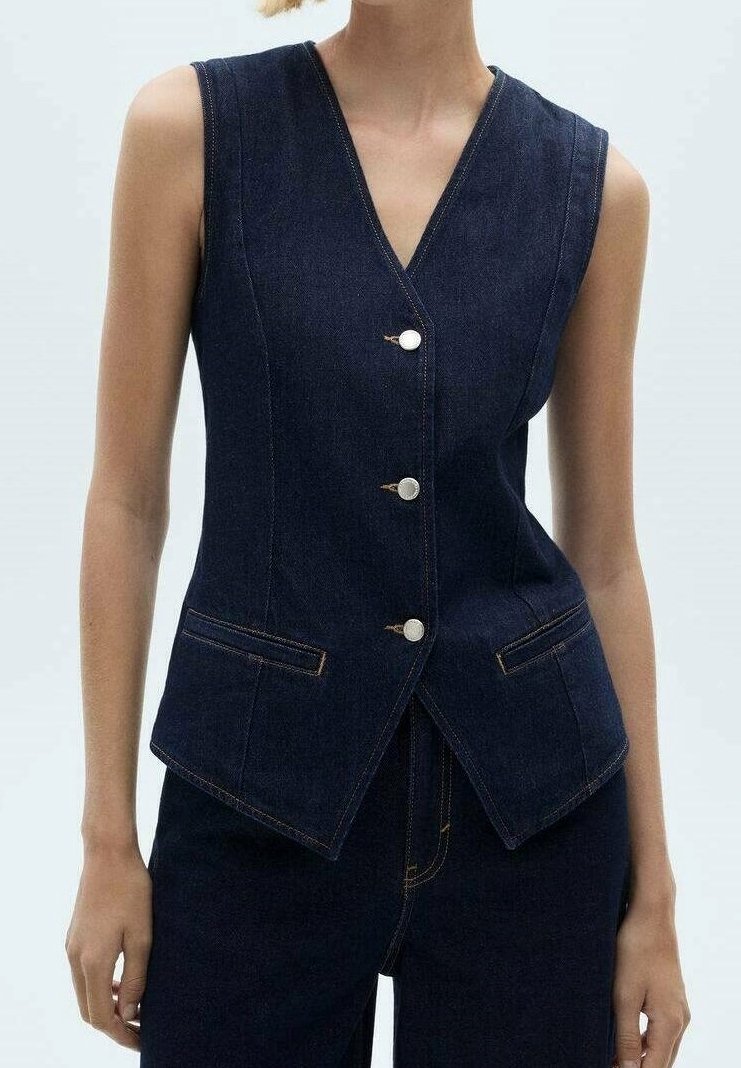 Veste en denim foncé sans manches avec col en V, trois boutons argentés et deux poches avant, portée avec un pantalon en denim foncé assorti.