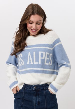 Frau lächelt nach unten, trägt einen weiß-hellblau gestreiften Pullover mit dem Schriftzug "LES ALPES" und eine dunkelblaue Jeans, Hände in den Taschen.
