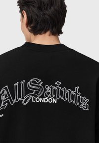 Felpa nera con testo ricamato bianco: "All Saints" in un font stilizzato con "LONDON" sotto. Design a girocollo.