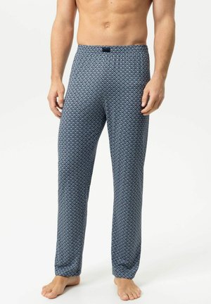 Mann trägt blaue gemusterte lange Pyjamahosen, steht barfuß vor einem schlichten hellen Hintergrund, Torso und Beine sichtbar.