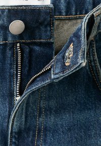Jeans de mezclilla azul oscuro con costuras visibles, un botón de metal y una cremallera plateada. Incluye una etiqueta en la cintura.