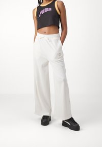 Osoba v černém zkráceném tank topu Puma, bílých širokých kalhotách s páskem na stažení a černých platformových teniskách stojí s rukama v kapsách.