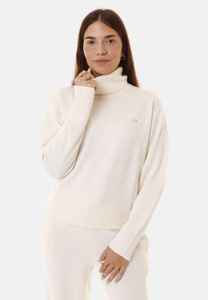 Maglione a collo alto crema con texture a costine e maniche lunghe, caratterizzato da una vestibilità leggermente cropped e un piccolo logo sul petto.
