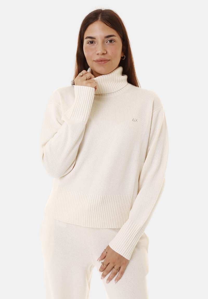 Maglione a collo alto crema con texture a costine e maniche lunghe, caratterizzato da una vestibilità leggermente cropped e un piccolo logo sul petto.