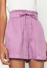 Lila bomullsshorts med elastisk midja, dragsko-detalj och sidofickor. Tyget har en slät yta och vikta fållar.