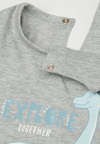 Šedá bavlněná košile s kulatým výstřihem, patentním zapínáním a modrou grafikou dinosaura s textem "PROZKOUMEJME SPOLEČNĚ." Hladká textura.