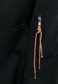 Ropa de tela negra con una cremallera plateada que está parcialmente bajada, con un cordón naranja y blanco pasado por el tirador de la cremallera.