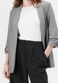 Blazer gris aux manches retroussées, associé à un t-shirt blanc et un pantalon noir taille haute avec plis sur le devant et poches latérales.