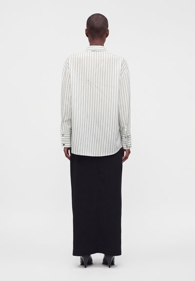 Personne portant une chemise blanche à manches longues avec de fines rayures noires et une longue jupe noire, vue de dos sur un fond uni.