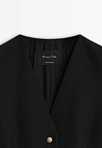 Vêtement noir avec un col en V profond, un bouton métallique unique et une étiquette intérieure indiquant "Massimo Dutti, Made in Morocco."