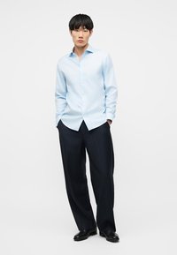 Νεαρός άνδρας όρθιος με τα χέρια στις τσέπες, φορά ανοιχτό μπλε πουκάμισο με κουμπιά, μαύρο παντελόνι με ρίγες ευρύ σε μήκος και μαύρα παπούτσια.