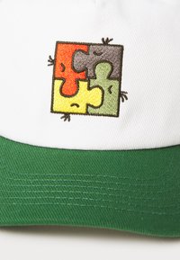 Hvit caps med grønn brem som har en brodert puslespillbit-design i rød, gul, grå og grønn, med små hånddetaljer.