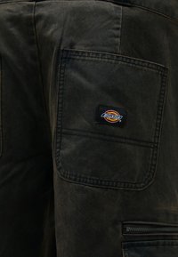 Σκούρα παντελόνια εργασίας από καμβά με τσέπη φθοράς και τσέπη με φερμουάρ, και μικρή ετικέτα λογότυπου στο πίσω μέρος. Υφασμάτινη υφή με ελαφρώς φθαρμένη εμφάνιση.