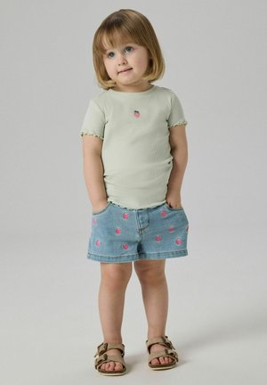 Young child standing with hands in pockets wearing a light green shirt and denim shorts with strawberry prints and brown sandals.