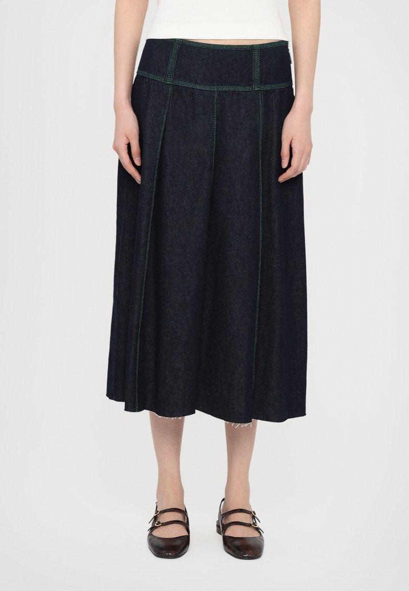 Jupe midi en denim foncé avec une large ceinture et des surpiqûres vertes, portée avec des chaussures plates noires à brides et un haut blanc.