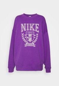 Lila tröja med rund halsringning i bomull, prydd med en stor vit Nike-logotyp, retro grafik från 1972 samt lagerbladselement vid halsringningen och fållen.