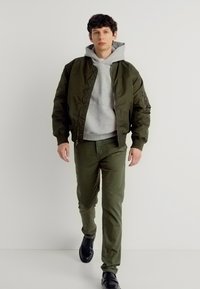 Νεαρός άνδρας που περπατά μπροστά φορώντας ένα μπουφάν μπόμβερ σε ελαιώδες πράσινο, ανοιχτό γκρι φούτερ, παντελόνι σε ελαιώδες πράσινο και μαύρα παπούτσια, με φόντο ένα απλό λευκό υπόβαθρο.