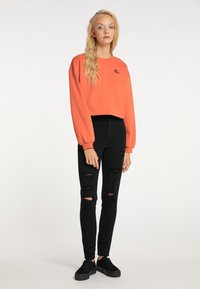 Tricou scurt portocalie cu mâneci lungi, asortat cu blugi skinny negri rupți și pantofi negri fara toc, stând în fața unui fundal simplu.