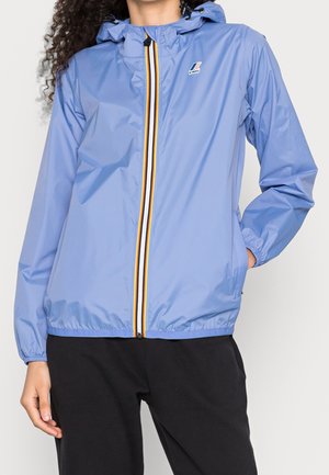 Person iført en lyseblå hættetrøje vindjakke med flerfarvet lynlås og sorte bukser, hånd i lommen, foran ensfarvet baggrund.