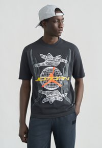 Μαύρο μπλουζάκι με γραφιστικό σχεδιασμό που απεικονίζει μια υδρόγειο σφαίρα, χέρια και μια κόκκινο-πορτοκαλί σιλουέτα παίκτη μπάσκετ. Συνοδεύεται από γκρι καπέλο.