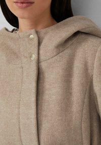 Manteau en laine beige avec une grande capuche, doté d'une finition texturée, d'un col arrondi et de boutons-pression dorés à l'avant.