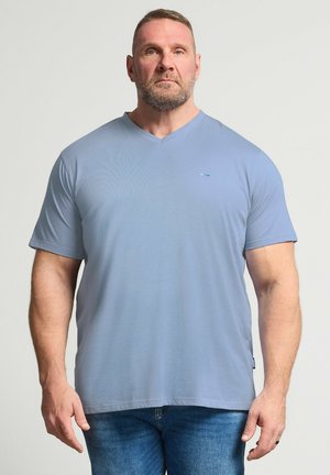 Uomo muscoloso con la barba che indossa una maglietta azzurro chiaro a maniche corte con scollo a V e jeans blu, in piedi davanti a uno sfondo chiaro uniforme.