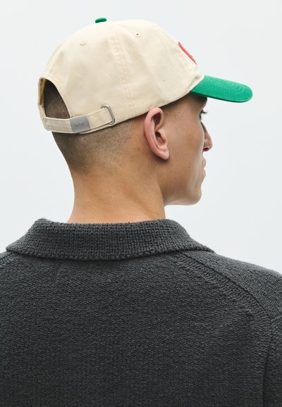 Side view of a person with short hair wearing a beige and green baseball cap and a dark textured sweater with a folded collar.