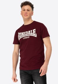 Burgundi színű pamut póló, nagy fehér "LONSDALE" logóval és ívelt dizájnú "LONDON" felirattal. Rövid ujjú és kerek nyakú.
