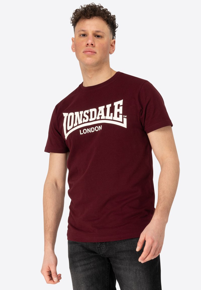 Bordo spalvos medvilnės marškinėliai su dideliu baltu "LONSDALE" logotipu ir kreivai užrašytu "LONDON" tekstu. Trumpomis rankovėmis ir apykakle.