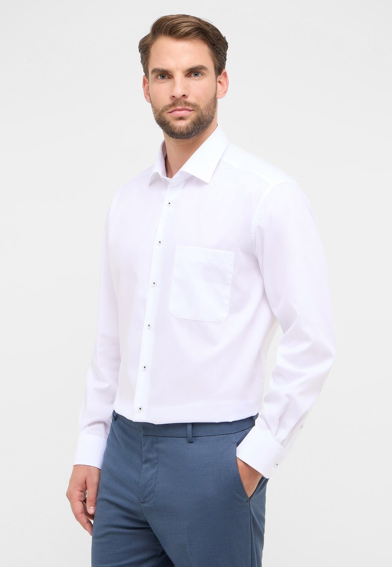 Camicia bianca a maniche lunghe con colletto alla francese, dotata di una tasca sul petto e bottoni in metallo, indossata con pantaloni blu.
