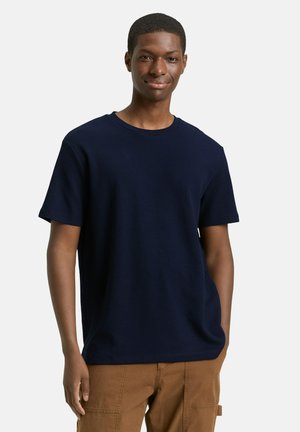 Joven con pelo corto, vestido con una camisa azul marino de manga corta y pantalones marrones, de pie y sonriendo con una mano en el bolsillo.