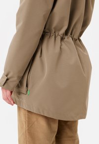 Giacca impermeabile color kaki con vita cinchata, texture liscia e polsini regolabili. Accentuata da un'etichetta verde vicino all'orlo.