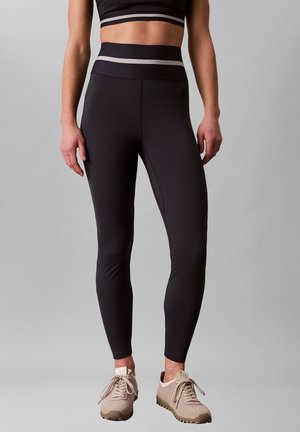 Femme portant des leggings Calvin Klein taille haute noirs, un soutien-gorge de sport noir et des baskets beige, debout devant un fond gris uni.