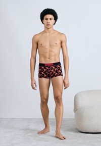 Chiloți boxeri negri cu un model floral roșu, având o talie roșie cu logo-ul "DIESEL", purtați de un model masculin în picioare.